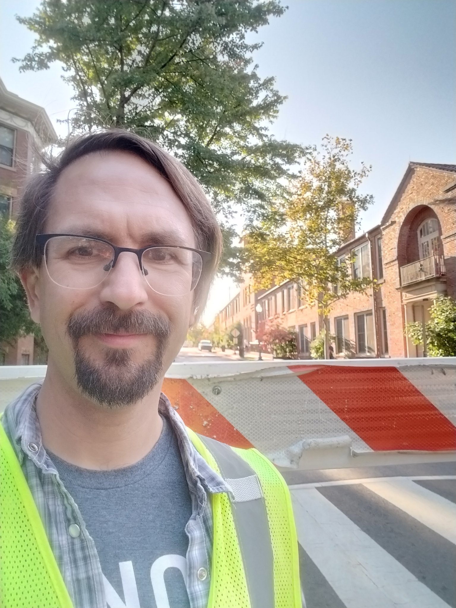 Photo of J. Swiderski, a white person with brown hair and goatee, wearing a yellow safety vest and standing in front of a traffic barrier outside Bancroft Elementary School in Mount Pleasant, DC.